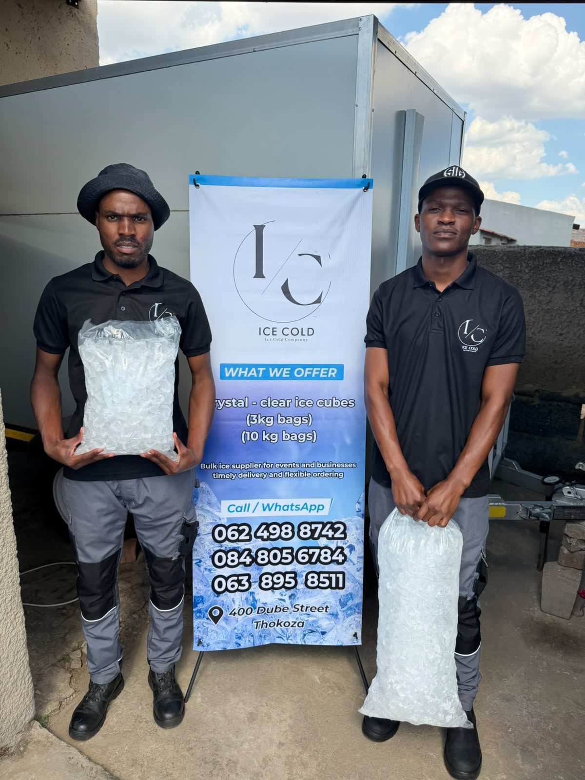 Two Ice Cold team members in branded shirts holding bags of ice beside the Ice Cold banner in Thokoza